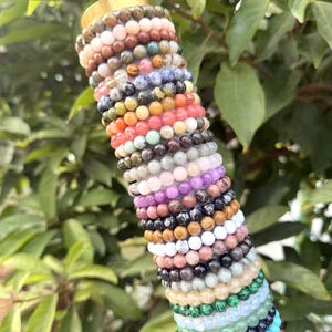 May include: A collection of beaded bracelets in various colors, including pink, orange, green, and black. The bracelets are stacked together, showcasing a variety of natural stone beads. The background features green foliage.