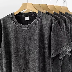 May include: Several dark gray, acid-wash style t-shirts hanging on wooden hangers. The shirts have a crew neck and short sleeves. The fabric has a textured appearance. The shirts are displayed against a white background.