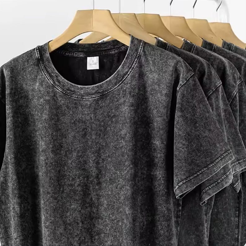 May include: Several dark gray, acid-wash style t-shirts hanging on wooden hangers. The shirts have a crew neck and short sleeves. The fabric has a textured appearance. The shirts are displayed against a white background.