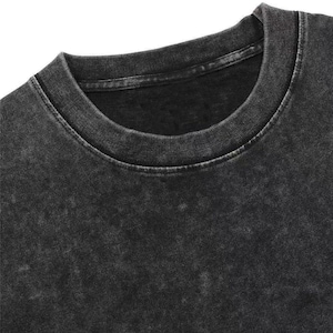 May include: Close-up of a black, acid-washed crew neck t-shirt. The shirt has a faded, textured appearance with a ribbed neckline. The fabric appears soft and worn, suggesting a vintage or retro style. The overall aesthetic is casual and comfortable.