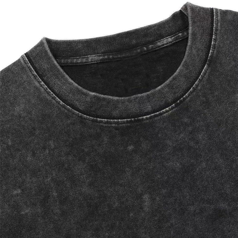 May include: Close-up of a black, acid-washed crew neck t-shirt. The shirt has a faded, textured appearance with a ribbed neckline. The fabric appears soft and worn, suggesting a vintage or retro style. The overall aesthetic is casual and comfortable.