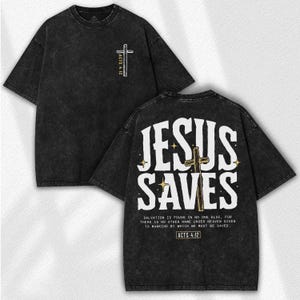 May include: Black, acid-wash style t-shirt with the text "JESUS SAVES" in white on the back, along with a cross graphic. The front features a smaller cross and "ACTS 4:12". The shirt includes a Bible verse.