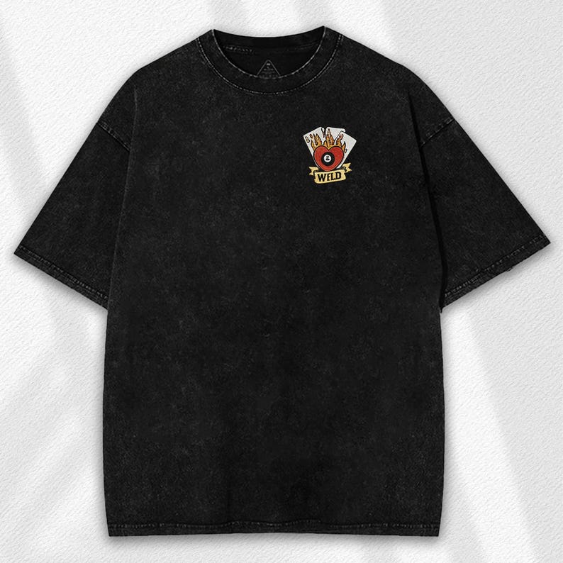 May include: Black, short-sleeved t-shirt with a faded, textured appearance. The shirt features a small graphic on the chest with a heart, a billiard ball, and playing cards, with the word "WILD" below. The shirt has a crew neck.