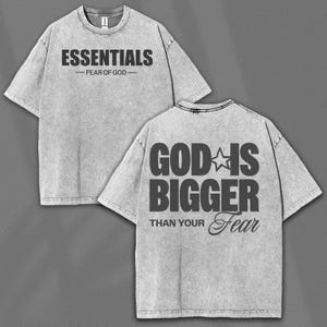 May include: Two gray t-shirts with black text. The front shirt reads "ESSENTIALS - FEAR OF GOD". The back shirt reads "GOD IS BIGGER THAN YOUR Fear" with a star graphic. The shirts have a textured, washed appearance.