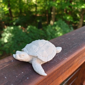 May include: A small, white, sculpted turtle figurine featuring a detailed, textured shell with geometric patterns and realistic scale markings on its head and flippers. The object is solid white with a matte finish, capturing the form of a sea turtle.