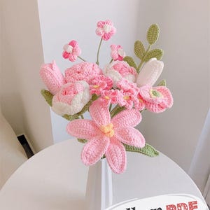 May include: A handmade crochet bouquet of pink and white flowers in a white vase. The bouquet includes various flower types, such as tulips and daisies, with green leaves. The vase sits on a white table. The text "Pattern PDF" is visible.