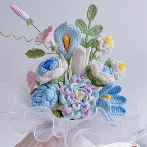 Könnte beinhalten: Ein handgefertigter Häkelblumenstrauß mit verschiedenen Blumen in Blau-, Weiß-, Rosa- und Lilatönen, mit grünen Blättern. Der Strauß ist in weißen Stoff gewickelt. Der Text "Pattern PDF" ist sichtbar.