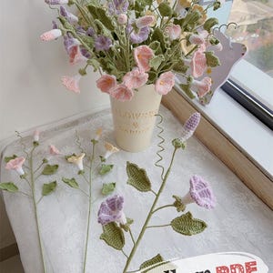 Könnte beinhalten: Ein Strauß handgehäkelter Blumen in einem cremefarbenen Metalleimer. Die Blumen sind in Rosa-, Lila-, Gelb- und Weißtönen gehalten, mit grünen Blättern. Auf dem Eimer steht "Flowers & Garden". Einzelne gehäkelte Blumenstiele sind ebenfalls zu sehen.