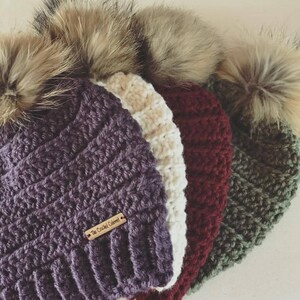 May include: Four crocheted beanies with faux fur pom poms. The beanies are in shades of purple, white, burgundy, and green. The purple beanie has a brown tag that reads "The Crochet Corner".
