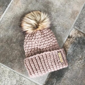 May include: A pink crocheted beanie with a fluffy brown pom pom and a small gold tag that says "Crochet Chic".