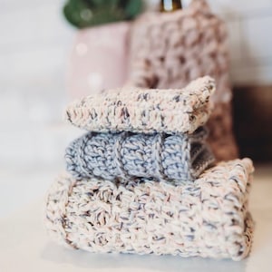 May include: A stack of three knitted dishcloths in various shades of pink, gray, and white. The dishcloths are made of a soft, absorbent material and are perfect for cleaning up spills and messes in the kitchen.