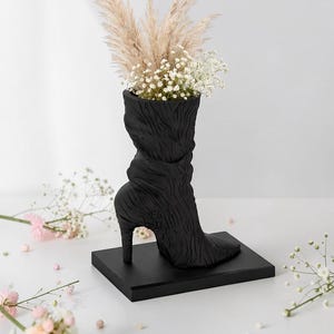 May include: A black, high-heeled boot-shaped vase with a textured surface, holding dried pampas grass and white flowers. The vase sits on a black rectangular base, creating a unique decorative piece. The overall theme is home decor.