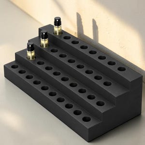 May include: A matte black tiered display stand with multiple circular cutouts for small cylindrical bottles. Three small clear glass spray bottles with black caps are inserted into the top and middle tiers of the rectangular organizer.