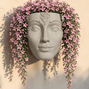 May include: A gray, wall-mounted planter shaped like a serene face, adorned with cascading pink flowers. The planter has detailed facial features and a decorative headpiece. The flowers create a flowing, floral 'hair' effect, adding a touch of elegance to the design.