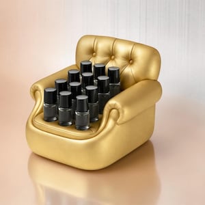 May include: A golden miniature armchair holds several black bottles with black caps. The chair has a tufted back and rolled arms. The background is a gradient of peach and white.