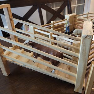 Peut inclure: Un ensemble de parc pour animaux de la ferme en bois avec une figurine de vache marron et blanche à l'intérieur. D'autres figurines d'animaux et une figurine d'agriculteur sont également visibles. Le parc est en bois clair.