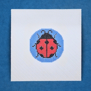 May include: A white needlepoint canvas featuring a detailed ladybug design. The ladybug is red with black spots and a black head, set against a light blue circle. The canvas is square, with a blue background.