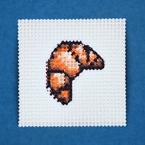 May include: A cross-stitch design of a croissant, featuring shades of orange, brown, and black. The pastry is set against a white, perforated background, with a blue backdrop. The image showcases a handmade food-themed craft.