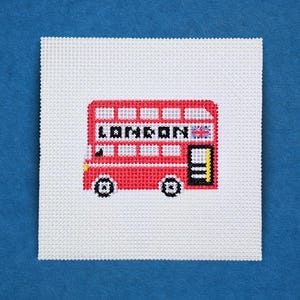 May include: A cross-stitch design featuring a red double-decker bus with the word "LONDON" and a Union Jack flag. The bus has black wheels and a yellow door. The design is on a white square canvas.