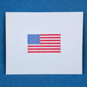 May include: A white square canvas with a cross-stitch American flag design. The flag features a blue canton with white stars and red and white stripes. The background is a solid blue.