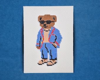 May include: Needlepoint art of a brown bear wearing sunglasses, a blue suit with red trim, and sandals. The bear is standing on a white background, with a blue background.
