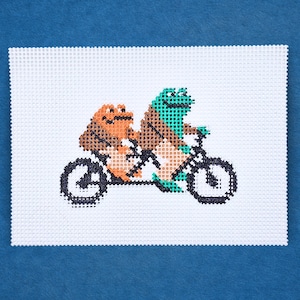 Puede incluir: Un diseño de punto de cruz con dos ranas montadas en un tándem. Una rana es naranja y marrón, la otra es verde y marrón. La bicicleta es negra y el fondo es blanco. La pieza está sobre un fondo azul.