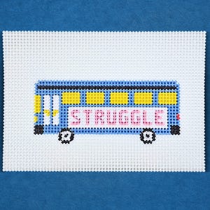 Könnte beinhalten: Eine Gobelin-Leinwand mit einem blauen Schulbus mit gelben Fenstern und dem Wort "STRUGGLE" in rosa Buchstaben. Der Bus hat schwarze Räder und ist vor einem weißen Hintergrund platziert.