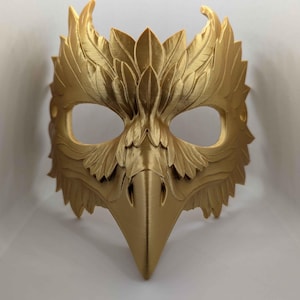 May include: A gold-colored mask with an avian design. The mask features a beak-like nose and is adorned with intricate feather and leaf details. The eye openings are oval-shaped. The mask is likely for costume or decorative purposes.