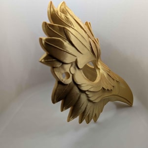 May include: A gold-colored, avian-themed mask with intricate feather detailing. The mask is designed to cover the upper face, featuring a beak-like structure and eye openings. The mask is likely made of a rigid material, and the design suggests a costume or decorative item.