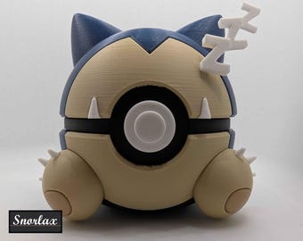 May include: A 3D-printed Snorlax figure in a sleeping pose, featuring a blue and tan color scheme with white accents. The figure has a round body, small arms, and a pair of Z's above its head. The word "Snorlax" is printed on a black rectangle.