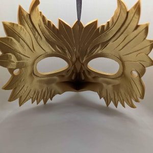 May include: A gold-colored, decorative mask with an intricate leaf design. The mask features eye cutouts and a detailed, textured surface. A black ribbon is attached for wearing. The mask is likely for costume or decorative purposes.