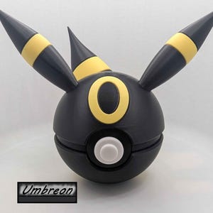 Może przedstawiać: Czarno-żółta figurka Umbreon, z kulistym korpusem z żółtym pierścieniem i akcentami. Posiada trzy spiczaste uszy z żółtymi paskami. Słowo "Umbreon" jest wyświetlane w prostokątnym polu na dole obrazu.