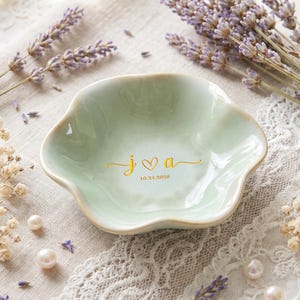 May include: A pale green ceramic trinket dish with a scalloped edge. The center features gold lettering with the lowercase letters j and a separated by a heart symbol, with the date 10.24.2026 printed in gold underneath the initials.