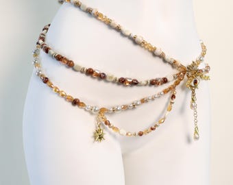 May include: A multi-strand beaded waist chain featuring cream, brown, and translucent amber-coloured beads. The design includes a central gold-toned eye charm with a dangling teardrop, a small star-shaped pendant, and various metallic accents.