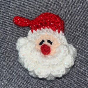 May include: A crocheted Santa Claus face with a red hat and nose, white beard, and black eyes. The handmade Christmas ornament is made with white and red yarn and is set against a grey background.