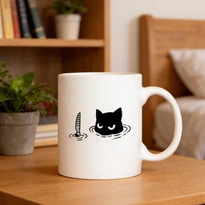 May include: White ceramic mug featuring a black cat head and a knife illustration. The cat's head is in water, and the knife is submerged. The mug has a curved handle and is on a wooden surface.