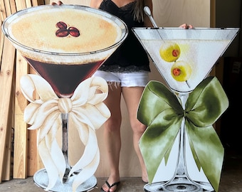 May include: Two large, flat, cutout-style cocktail glass decorations. The left glass contains a dark espresso martini topped with three coffee beans and a large cream-colored bow. The right glass holds a clear martini with two olives and a large olive-green bow.