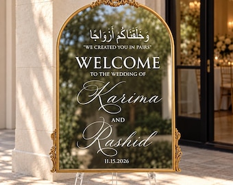 May include: An arched mirror wedding welcome sign with a gold frame featuring ornate detailing at the top and sides. The text reads: "وَخَلَقْنَاكُمْ أَزْوَاجًا", "WE CREATED YOU IN PAIRS", "WELCOME TO THE WEDDING OF Karima AND Rashid 11.15.2026".