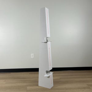 May include: A tall, white, multi-tiered cardboard floor display stand with a tapered, vertical structure. The unit features two distinct horizontal shelves or cutouts for product placement, with thin red vertical text printed along the front edges.