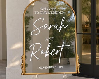 May include: An arched mirror wedding sign with a gold frame featuring ornate detailing at the top and sides. The white text reads: WELCOME TO OUR WEDDING, Sarah AND Robert, NOVEMBER 7, 2026. The mirror is displayed on a clear acrylic easel stand.