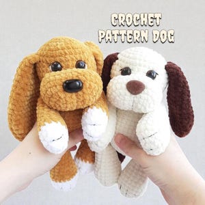 Peut inclure: Deux peluches de chien au crochet. L'une est beige avec des accents blancs et un nez noir. L'autre est blanche avec des accents marron. Le texte "CROCHET PATTERN DOG" est au-dessus des chiens.