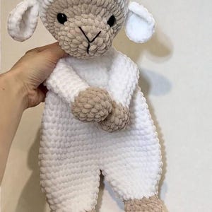 May include: A handmade crochet lamb toy. The lamb has a white body, head, and ears, with beige accents on the face, hands, and feet. It has black eyes and a stitched mouth. The lamb is soft and plush.