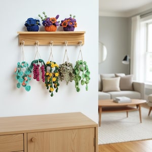 Puede incluir: Una estantería de madera exhibe tres arreglos florales de ganchillo en azul, naranja y morado. Debajo, seis plantas colgantes de ganchillo en varios colores, incluyendo turquesa, verde y amarillo, están suspendidas de la estantería con hilo.