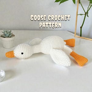 May include: A white crochet goose toy with orange feet and beak, lying on a white surface. The text "GOOSE CROCHET PATTERN" is displayed above the goose. A small succulent plant in a white pot is in the background.