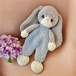 Op de afbeelding: Een handgemaakt grijs en blauw gehaakt konijn knuffel met lange oren, blauwe ogen en een roze neus. Het konijn draagt een blauwe jumpsuit en heeft witte accenten op zijn poten en voeten. Een boeket paarse bloemen staat op de achtergrond.