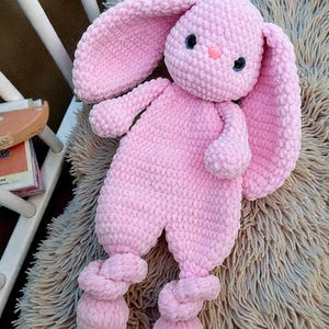 May include: A handmade pink crochet bunny plush toy with long floppy ears, blue eyes, and a pink nose. The bunny is lying on a fluffy, light brown surface. The bunny's arms and legs are attached to the body.