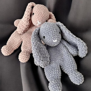 Puede incluir: Dos juguetes de peluche con forma de conejito en colores pastel suaves. Uno es rosa claro, el otro gris. Ambos tienen orejas largas y caídas, ojos negros pequeños y detalles blancos. Los conejitos están sobre una superficie gris oscuro.