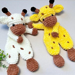 May include: Two handmade crochet giraffe plush toys. One is white with brown spots and accents, the other is yellow with brown spots and accents. Both have brown faces, ears, and feet. The giraffes are lying down.