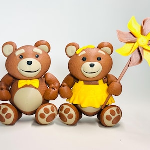 May include: Two brown teddy bear figurines. One bear wears a yellow bow tie, the other a yellow dress. The second bear holds a pinwheel with yellow and brown blades. The bears have light brown paws and ears.