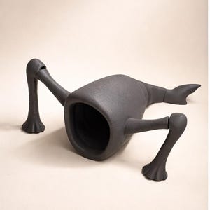 May include: A dark gray, abstract sculpture resembling a creature with a large opening and multiple appendages. The sculpture has a smooth, matte finish and is positioned on a neutral background.